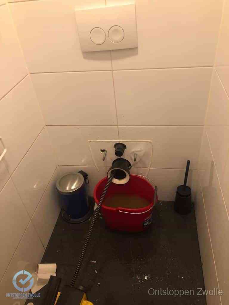 Een ontstopte wand-wc met verwijderde toiletpot waar een ontstoppingsveer in de wanduitgang verdwijnt, het achterhalen waar de verstopping zit in Zwolle