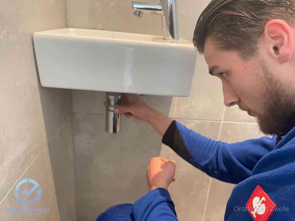 Een monteur draait met de hand de zwanenhalssifon onder een fonteinwasbak in de toiletruimte los om de bron van rioollucht in Zwolle te vinden