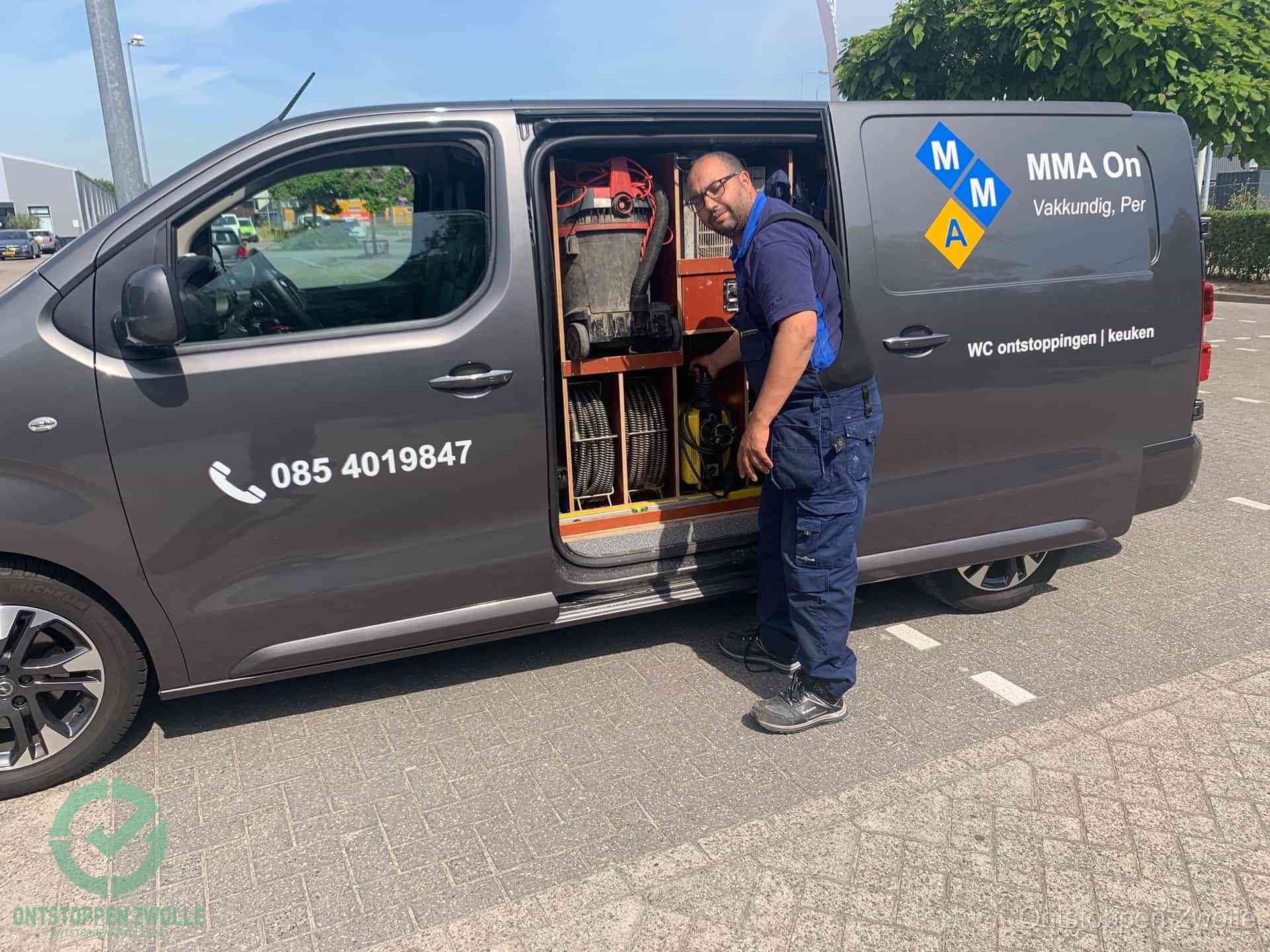Een monteur bij de open schuifdeur van zijn bedrijfsbus op een parkeerplaats, klaar om de oorzaak van terugkerende afvoerverstoppingen aan te pakken in Zwolle